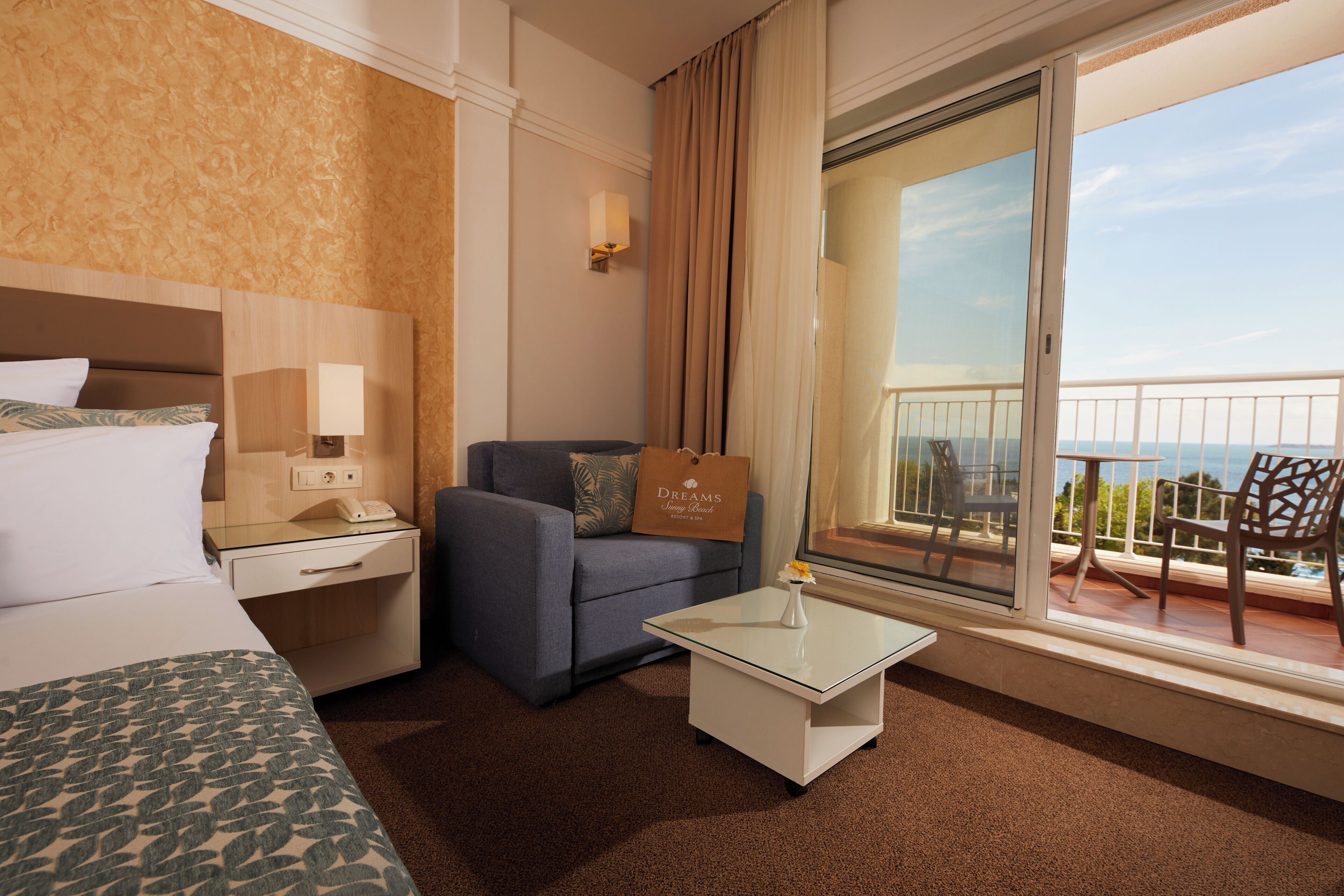 A hotel room with a bed, a sofa, a small table, a nightstand, a window with a balcony, and a view of the sea.