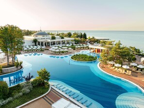A resort pool with sun loungers and umbrellas, surrounded by greenery and a view of the sea.
