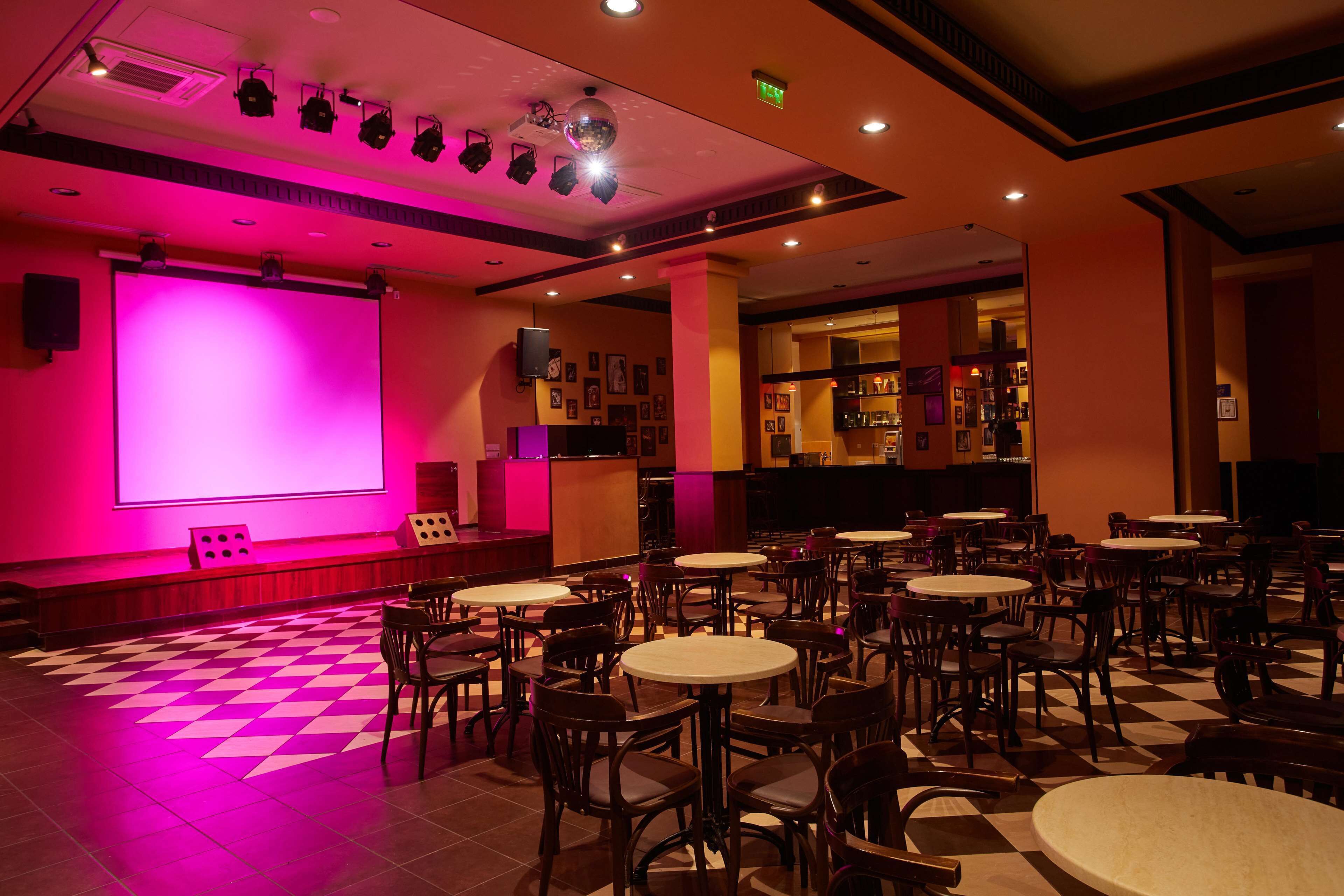 A restaurant with a checkered floor, multiple tables and chairs, a bar area, and a large screen on the wall.