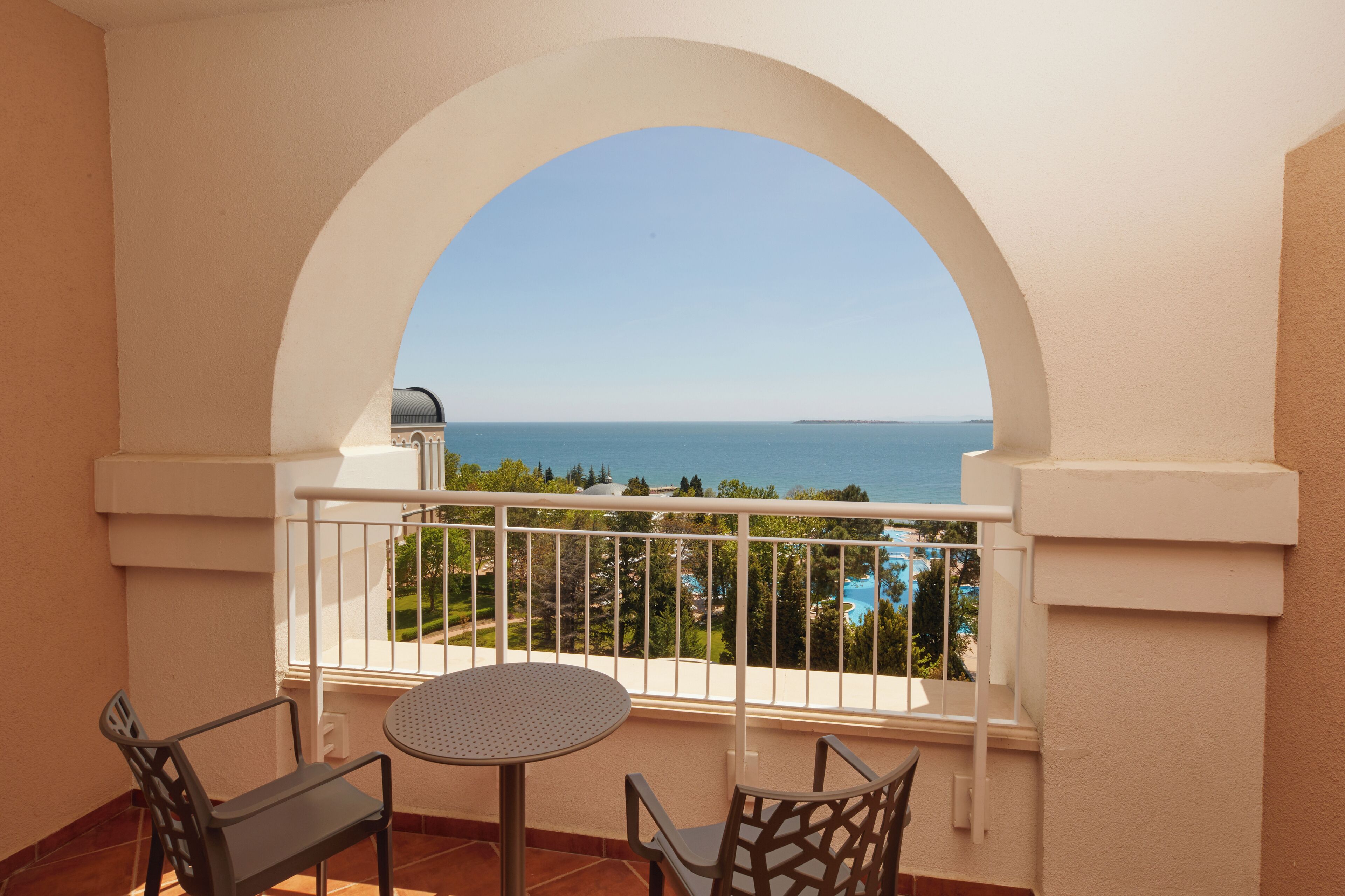 A balcony with a table and chairs, overlooking a clear blue sea and a distant shoreline.