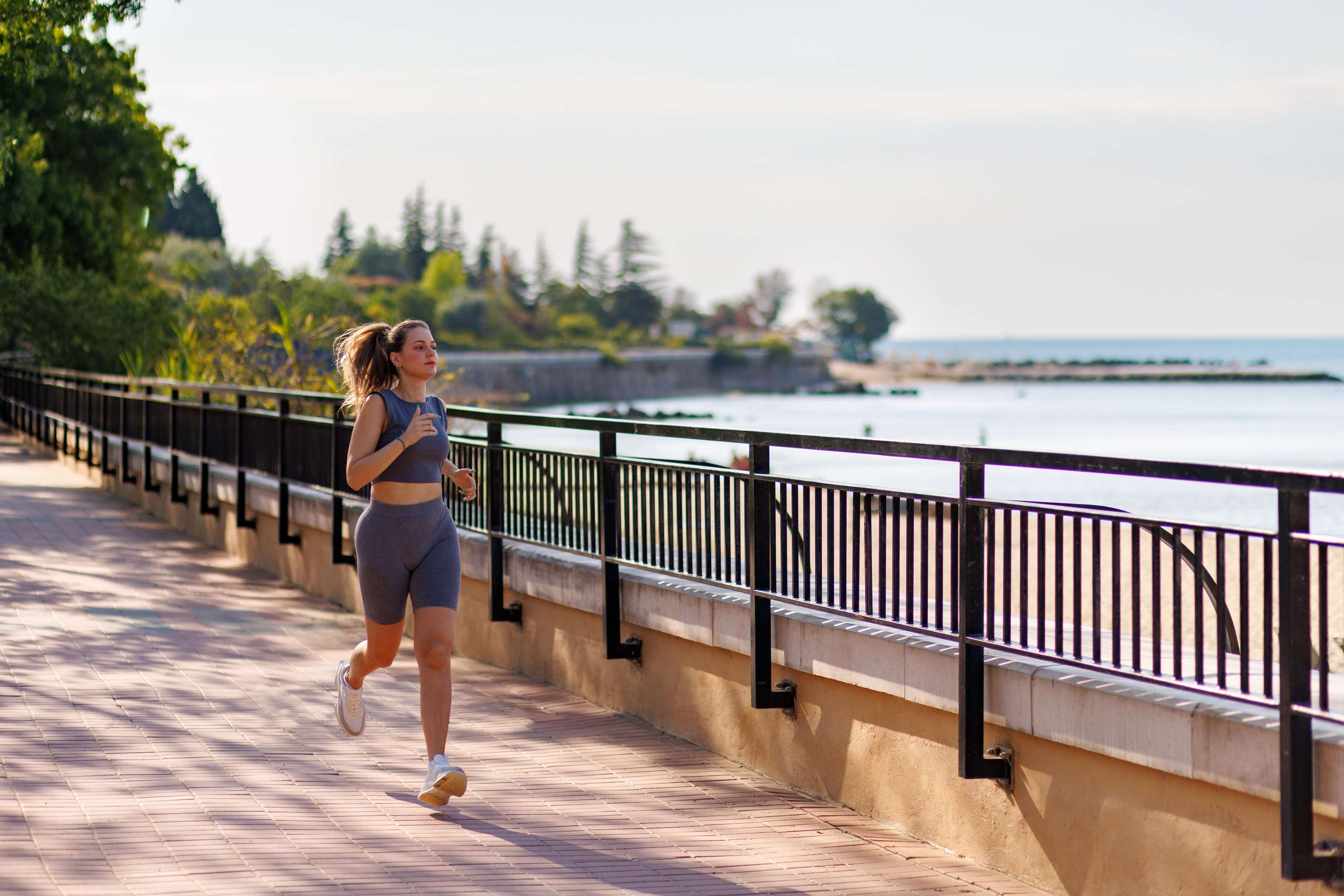 A woman jogging on a paved path beside a body of water, with a railing and greenery in the background.