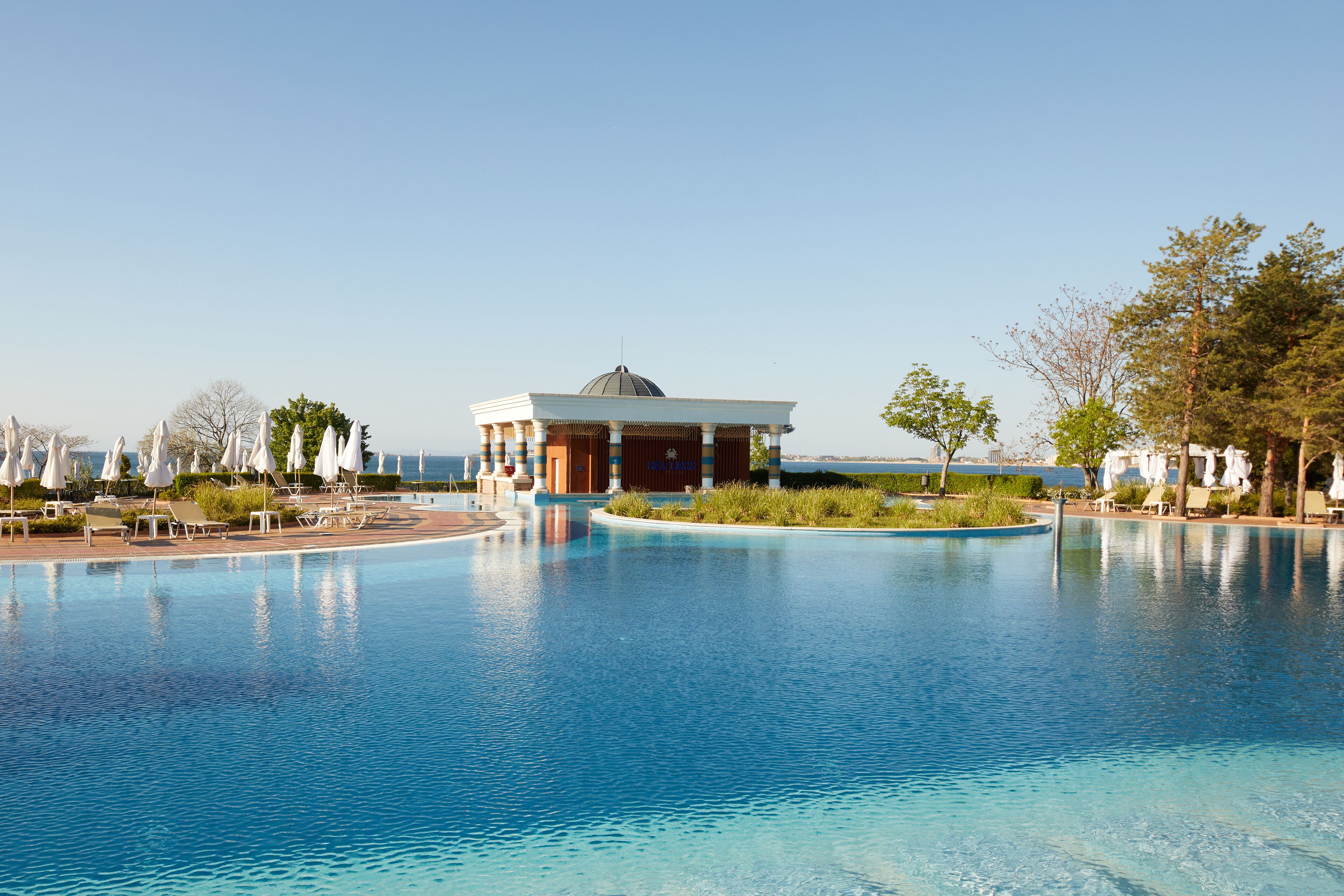 A swimming pool with lounge chairs and umbrellas, a building with a dome, and a view of the sea.