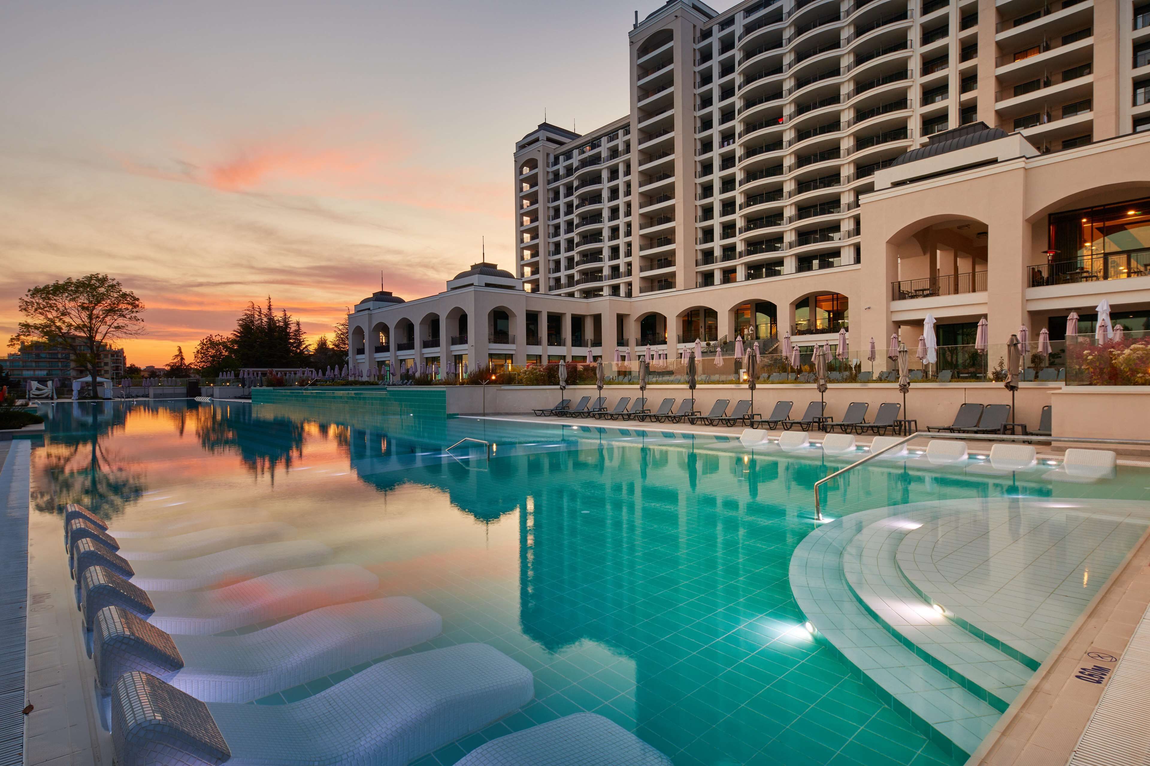 Un hotel con una piscina grande, sillas de descanso y vistas al atardecer.