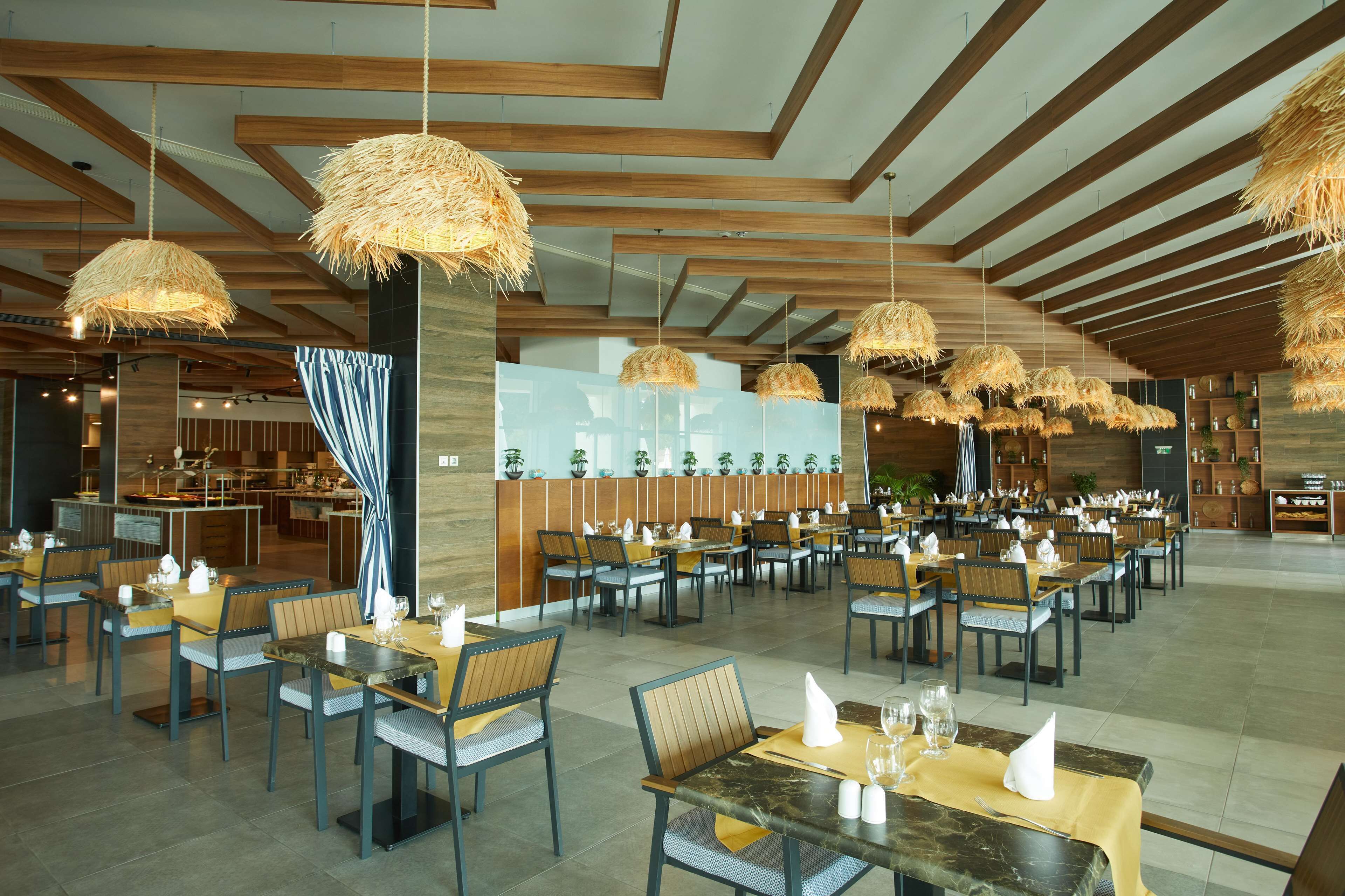 A spacious restaurant with wooden beams and hanging thatch lamps, tables set with yellow tablecloths and chairs.