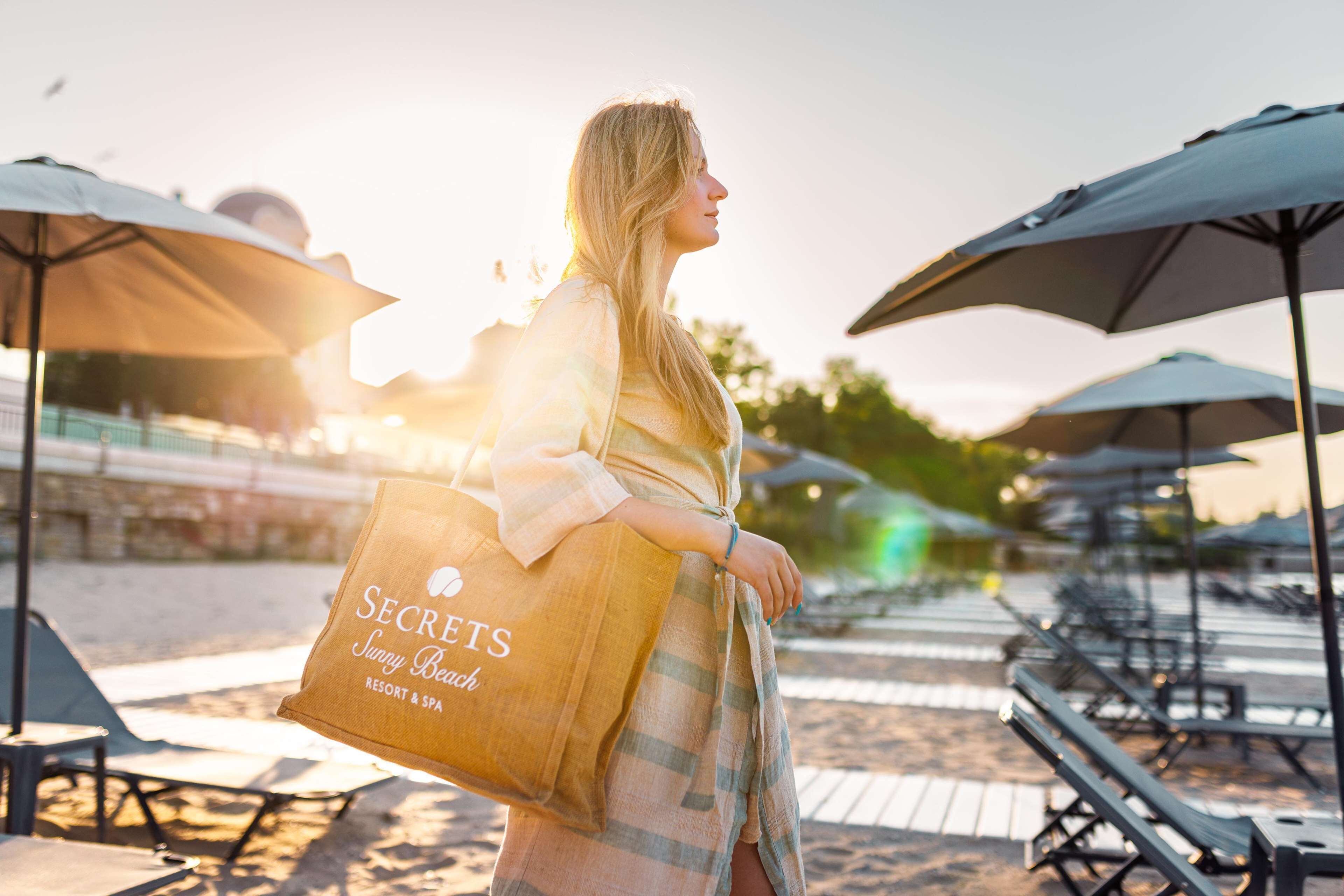 A person holding a bag with "SECRETS Sunny Beach Resort & Spa" text, walking on a beach with umbrellas and lounge chairs.