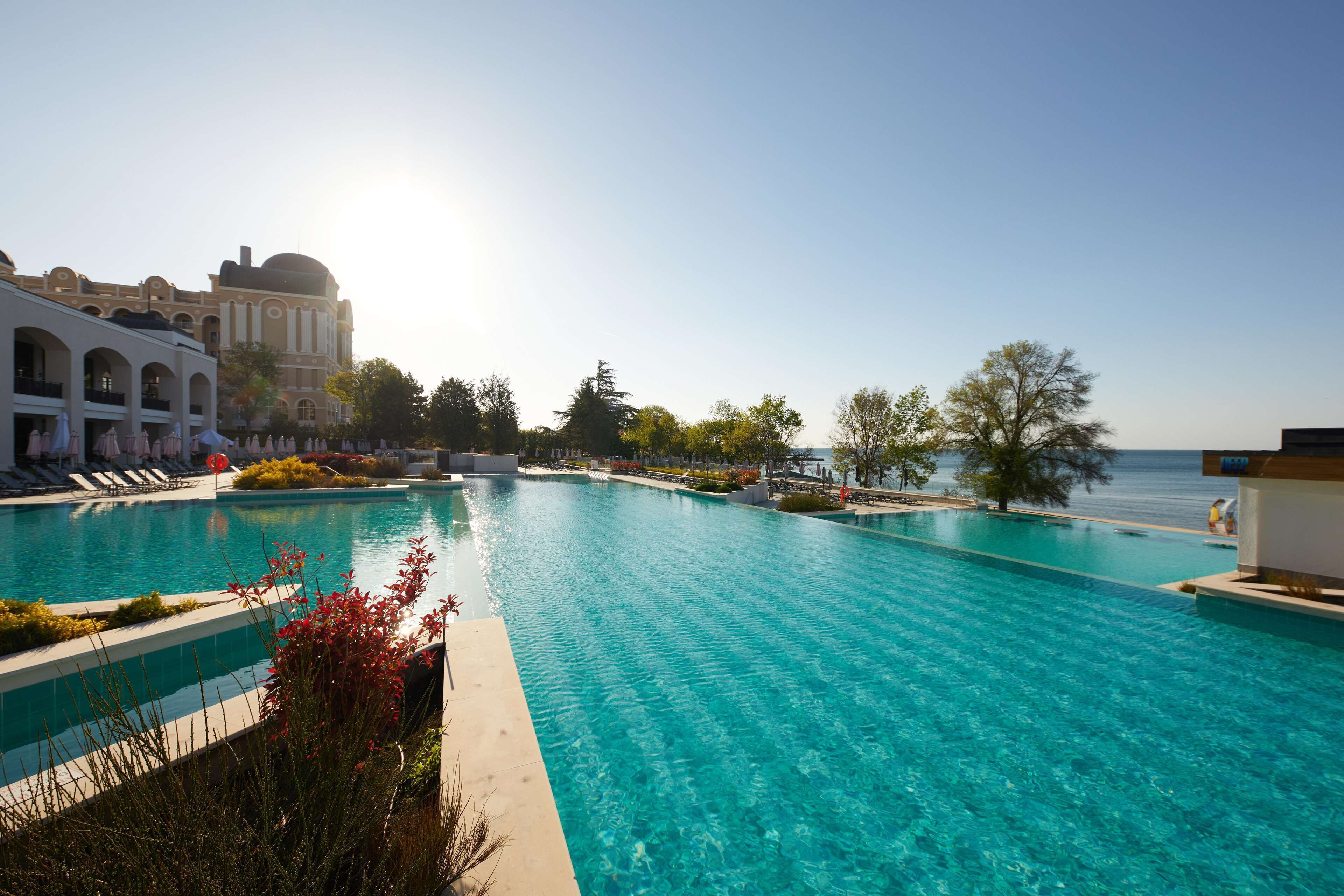 Une grande piscine extérieure à l’eau d’un bleu limpide, entourée de chaises longues et bordée d’un bâtiment aux fenêtres en arc de cercle.