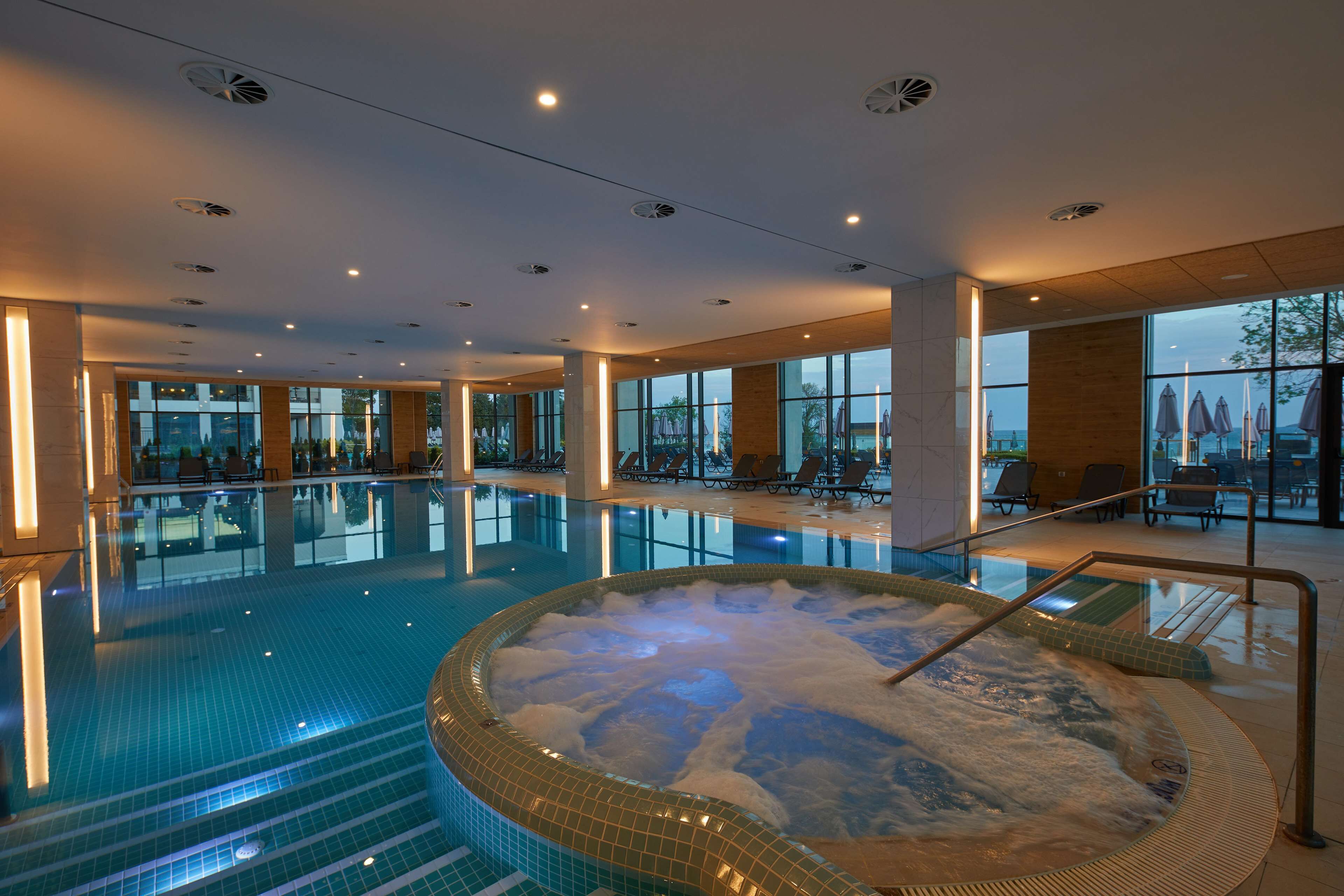 A large indoor swimming pool with a hot tub, lounge chairs, and a view of the outdoors.