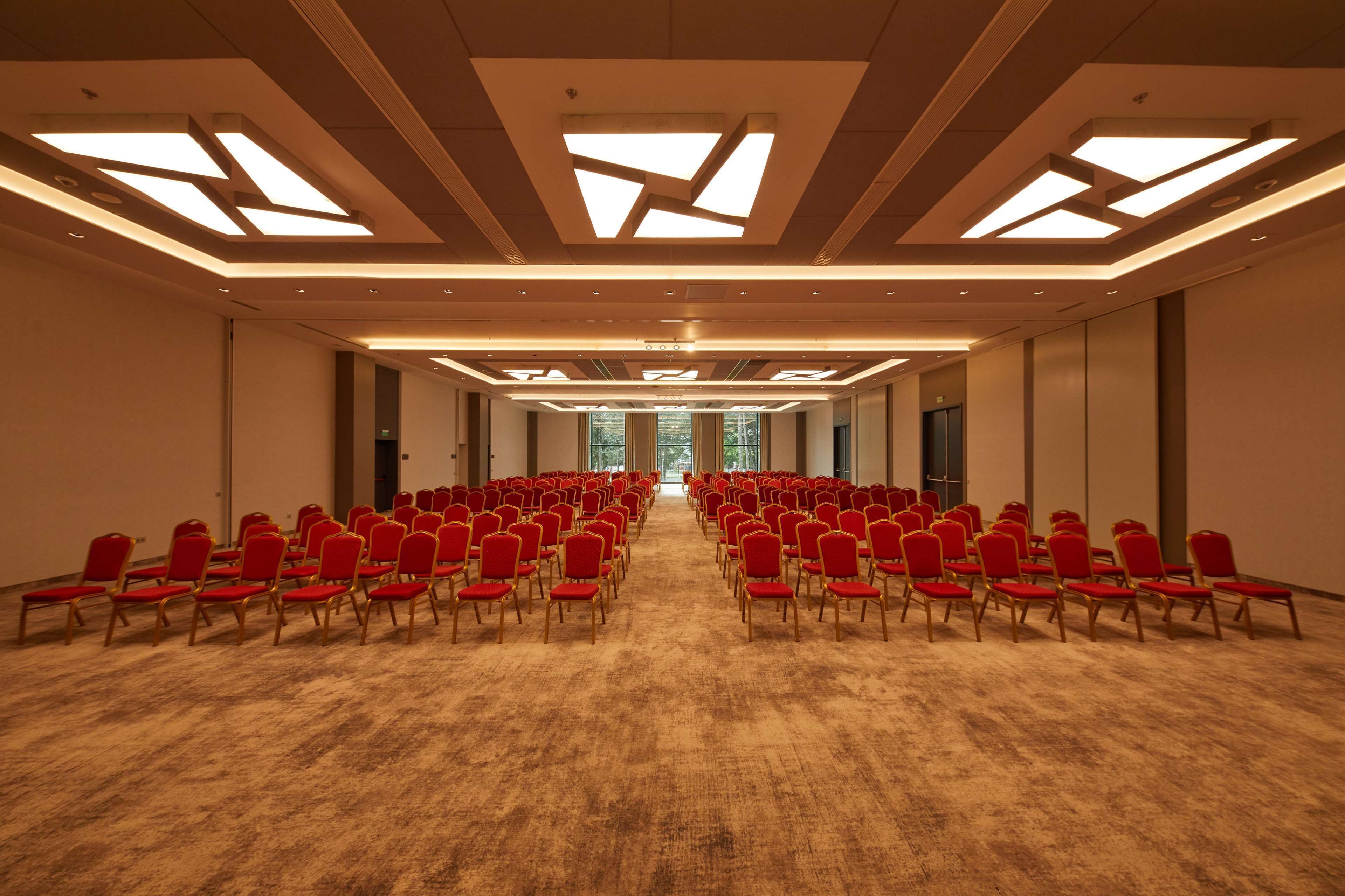 Een vergaderzaal met rijen rode stoelen, een vloerbedekking en een modern plafondontwerp met inbouwverlichting.