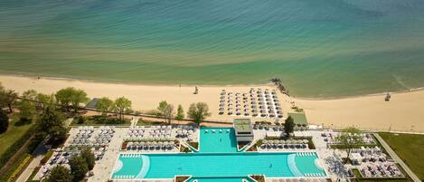 An aerial view of a resort with a large swimming pool, beach area, and numerous umbrellas.