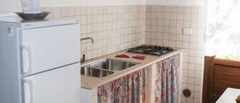 Una cocina compacta con un refrigerador blanco, una estufa a gas y un lavamanos. Un candelabro cuelga sobre la estufa, y la cocina tiene un salpicadero de azulejos.