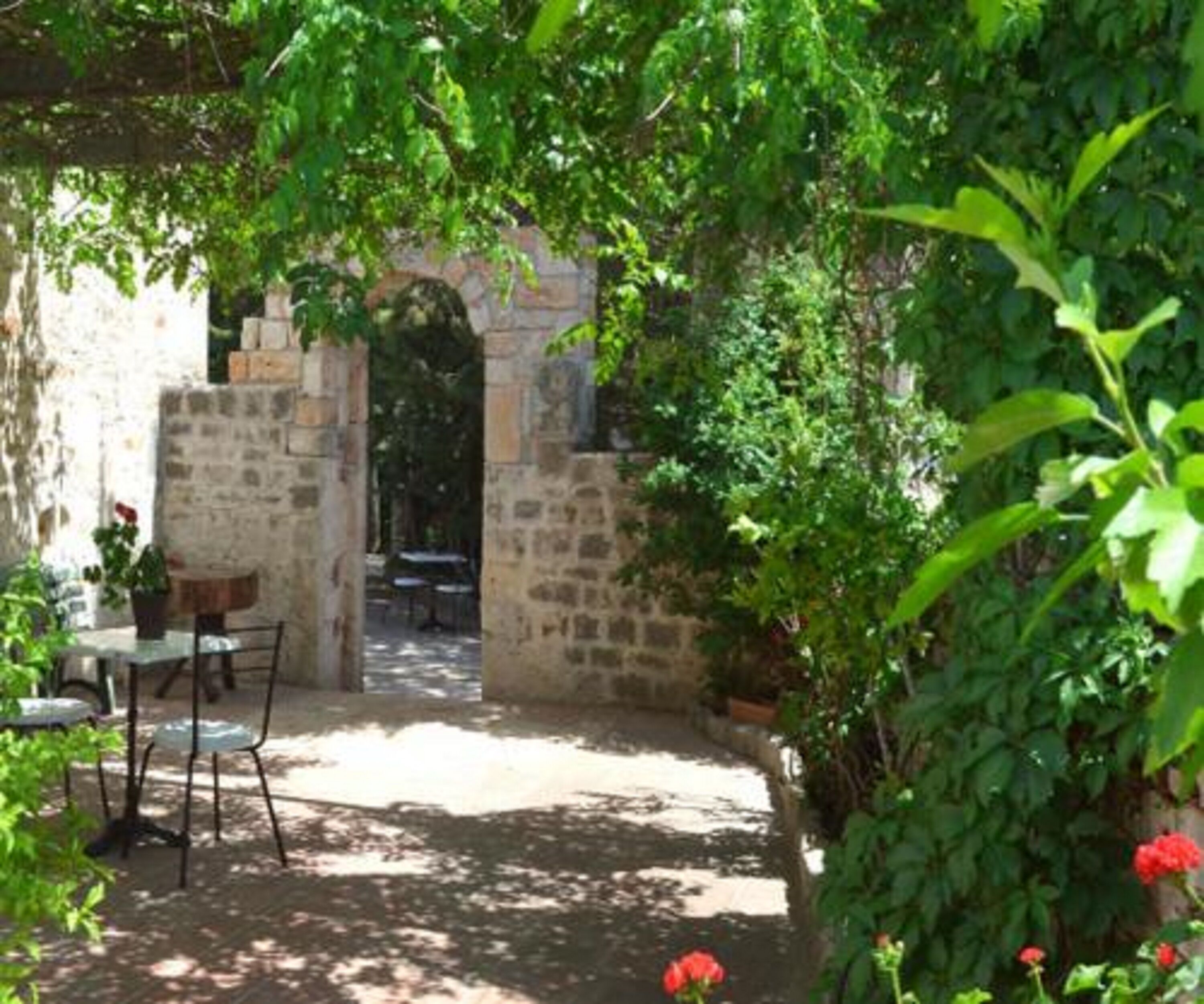 A stone courtyard with a table and chairs, surrounded by lush greenery and a stone wall.