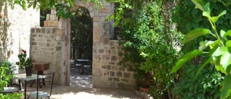 A stone courtyard with a table and chairs, surrounded by lush greenery and a stone wall.