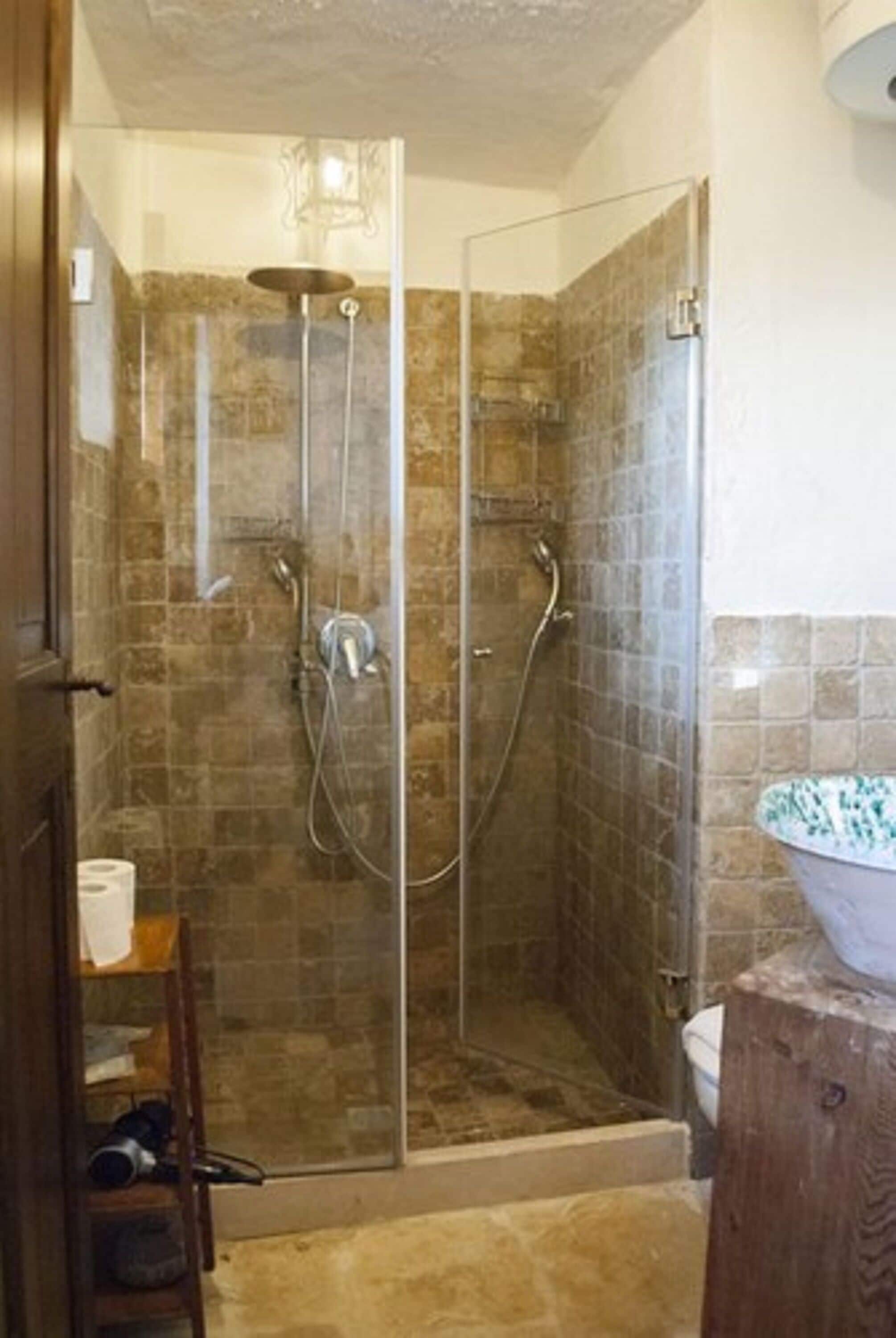 A glass-enclosed shower with a stone-tiled wall, a wooden door, and a white basin on a wooden stool.