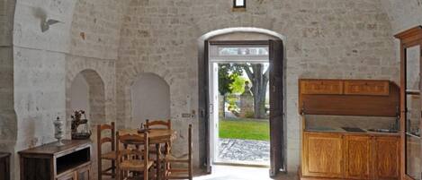 A rustic stone-walled room with a wooden dining table and chairs, a wooden cabinet, and a small window.