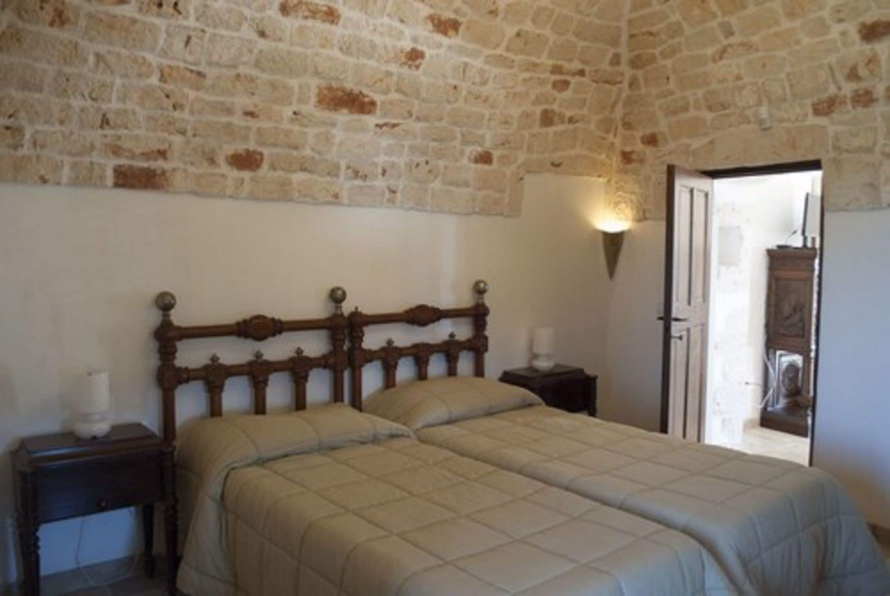 A bedroom with a brick wall, a wooden bed frame, a bedside table with a lamp, and a door leading to another room.