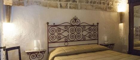 A bedroom with a stone wall, a metal bed frame, a bedside table with a lamp, and a chair.