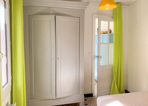 A room with a white wardrobe, a bed with a white bedspread, and a window with a view of a blue building. - Homerez - Charming House Soulac-sur-Mer, Garden, 65 m² (Soulac-sur-Mer)