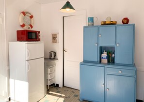 A compact kitchen with a white refrigerator, a blue cabinet with drawers, a red microwave, and a hanging light fixture. - Homerez - Charming House Soulac-sur-Mer, Garden, 65 m² (Soulac-sur-Mer)