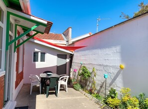 A patio area with a table and chairs, a white wall, and a garden with various plants and flowers. - Homerez - Charming House Soulac-sur-Mer, Garden, 65 m² (Soulac-sur-Mer)