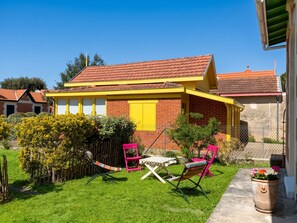 A house with a red-tiled roof, yellow trim, and a brick facade. There is a small garden with grass, a fence, and two pink chairs. - Homerez - Charming House Soulac-sur-Mer, Garden, 65 m² (Soulac-sur-Mer)