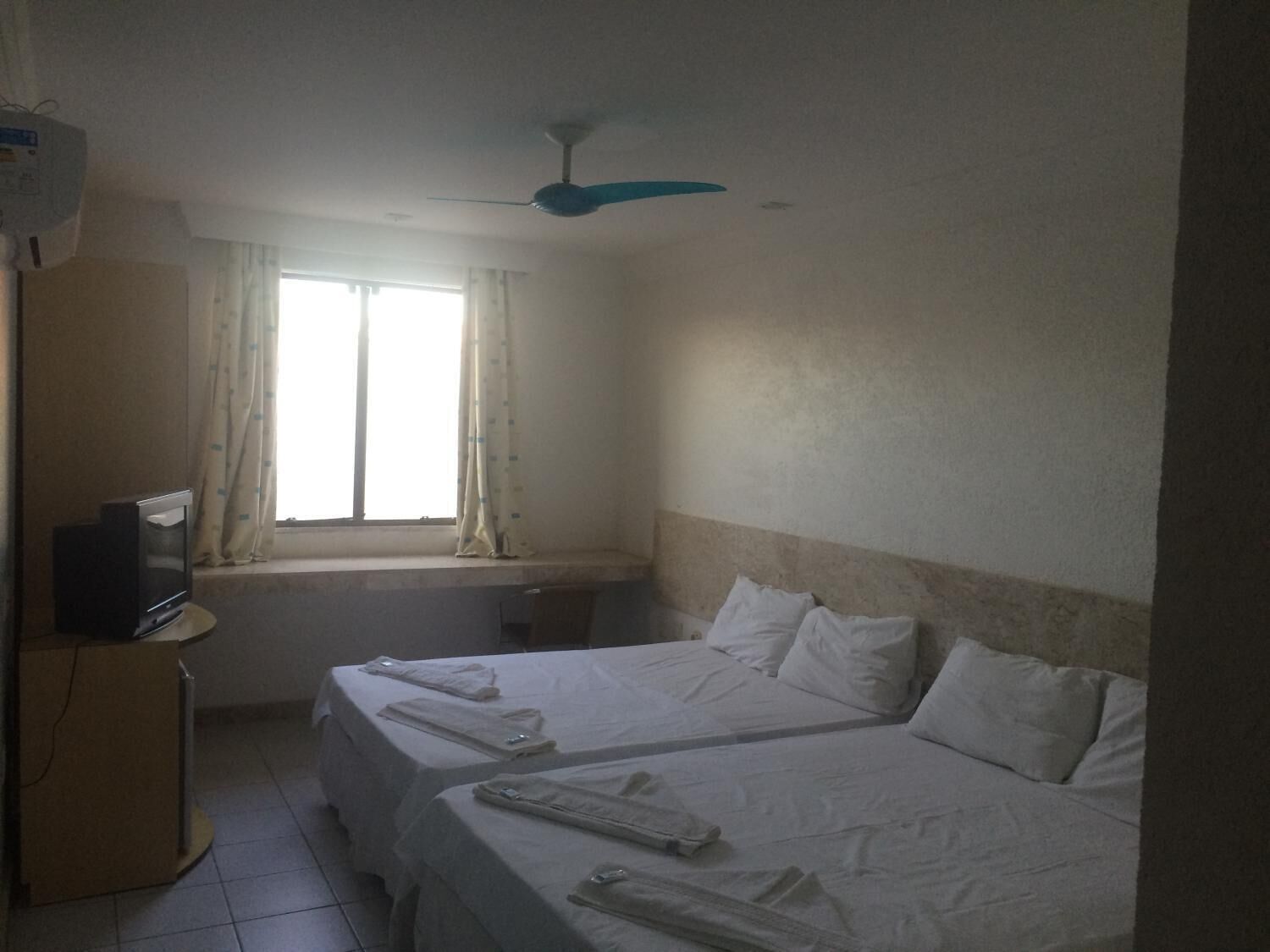 Habitación de hotel con dos camas, un televisor sobre un mueble, un ventilador de techo y una ventana con cortinas.