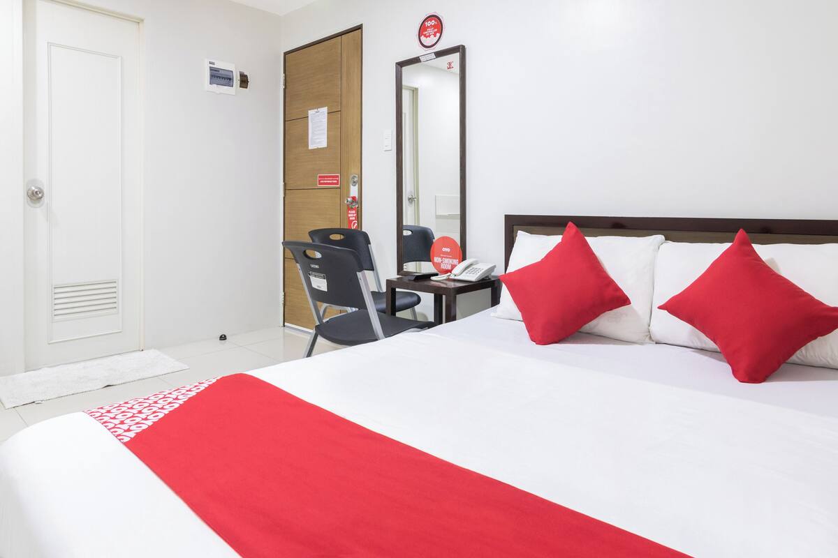A hotel room with a bed, two red pillows, a desk with a phone, and a mirror.