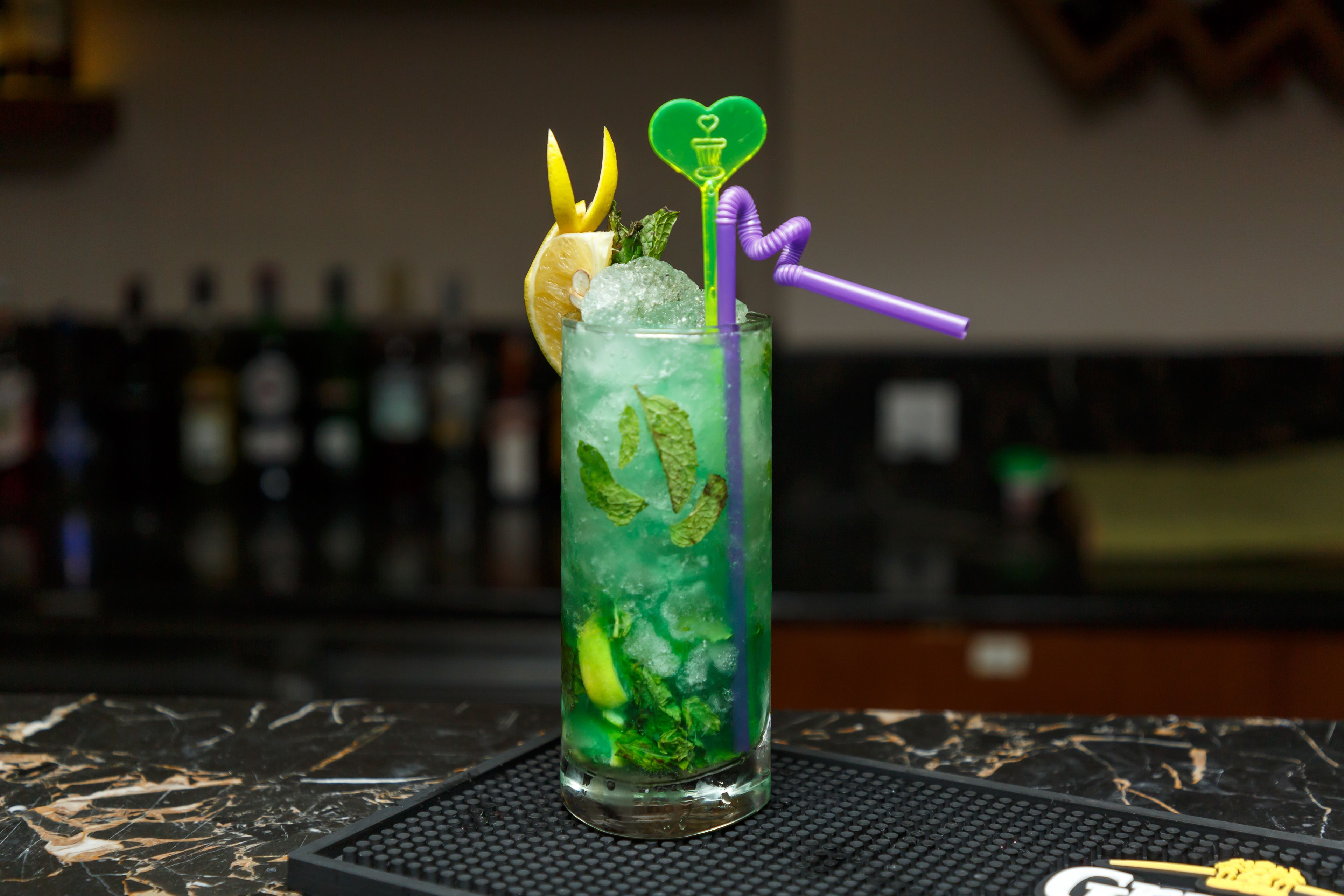 Un cocktail agrémenté d’une tranche de citron vert, de feuilles de menthe et d’une décoration en forme de cœur, le tout posé sur un tapis de bar noir.