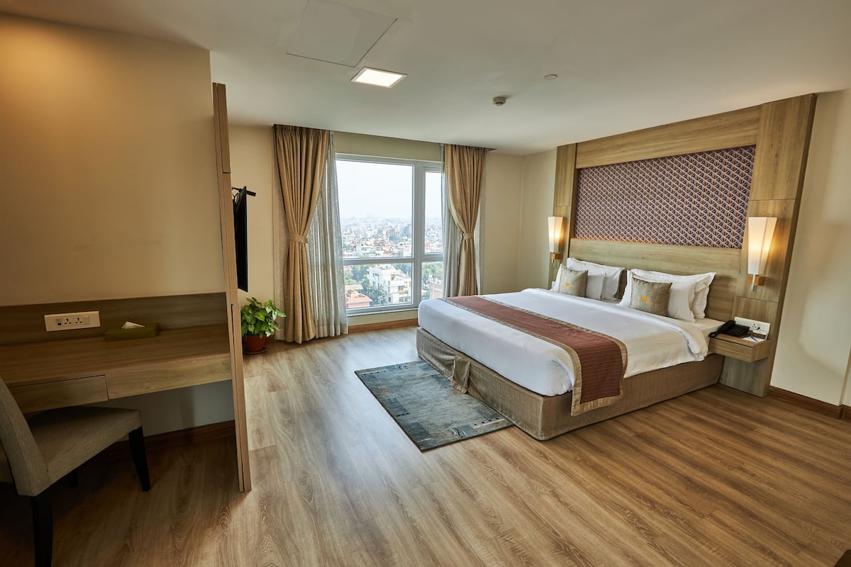 A hotel room with a large bed, a desk, a chair, a window with curtains, and a view of the city.