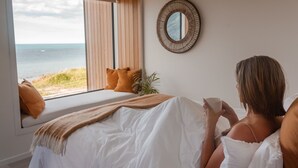 Un dormitorio con un ventanal grande con vistas a la playa, una cama con ropa blanca y un espejo redondo en la pared.