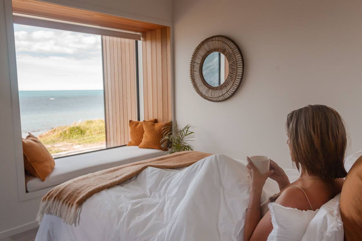 A bedroom with a large window overlooking the beach, a bed with white bedding, and a round mirror on the wall.