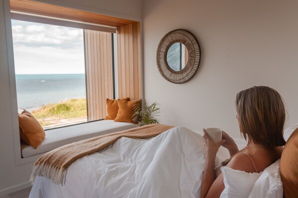 A bedroom with a large window overlooking the beach, a bed with white bedding, and a round mirror on the wall.