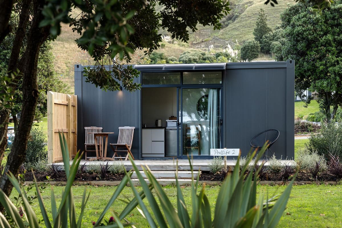 A modern, compact building with a sliding glass door entrance, a small outdoor seating area with two wooden chairs, and a sign indicating "Podlite 2".