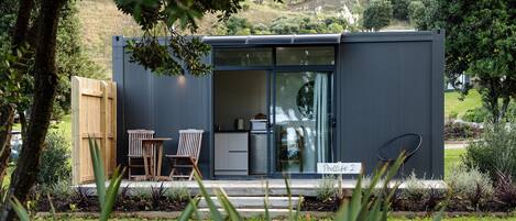 A modern, compact building with a sliding glass door entrance, a small outdoor seating area with two wooden chairs, and a sign indicating "Podlite 2".