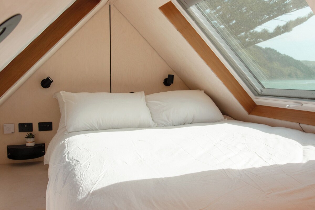 Een slaapkamer met een schuine plafond, een bed met wit beddengoed, een klein nachtkastje met een lamp, en een raam met uitzicht op een bebost gebied.