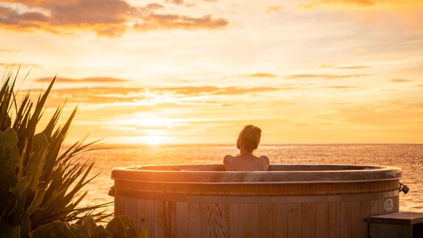 Una persona relajándose en un jacuzzi de madera con vista al atardecer.