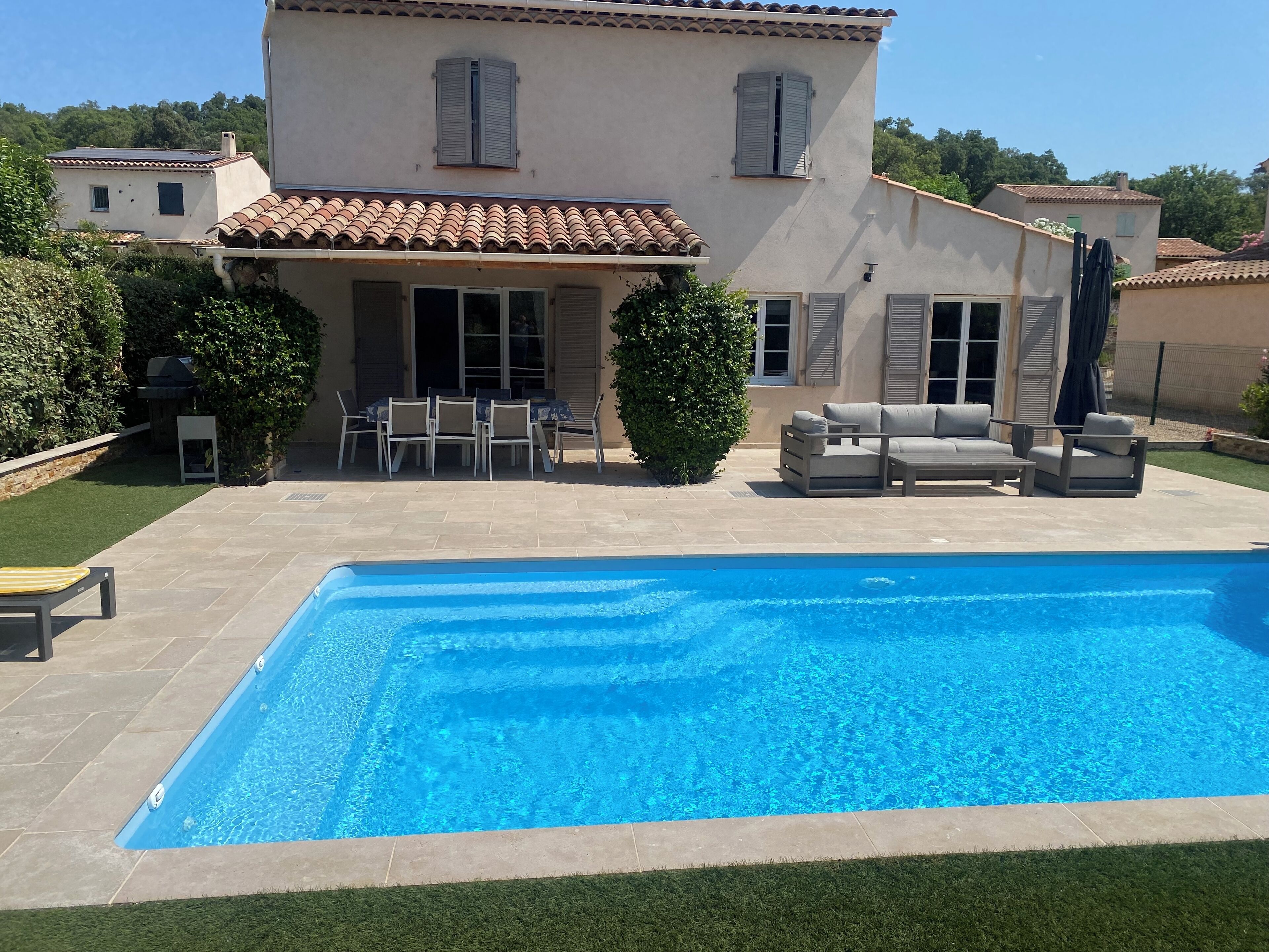 En swimmingpool med flisebelagte omgivelser, en terrasse med udendørs møbler og en bygning med flisebelagt tag og skodder.