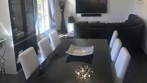 Ein moderner Essbereich mit einem schwarzen Tisch, weißen Stühlen und einer Kristallleuchter. Ein Flachbildfernseher ist an der Wand über dem Tisch angebracht.