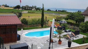 A swimming pool with a sun lounger and a covered seating area, surrounded by a fenced yard with a view of the countryside. - Chez Minnie (La Grande-Béroche)
