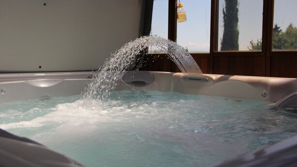 A hot tub with water cascading into it, set against a backdrop of wooden walls and a window. - Chez Minnie (La Grande-Béroche)