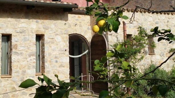 Un bâtiment rustique doté d’une tour rose et de murs en pierre, avec un patio agrémenté de chaises et d’une table en métal, le tout entouré de verdure et de citronniers.