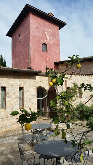 Un bâtiment rustique doté d’une tour rose et de murs en pierre, avec un patio agrémenté de chaises et d’une table en métal, le tout entouré de verdure et de citronniers.
