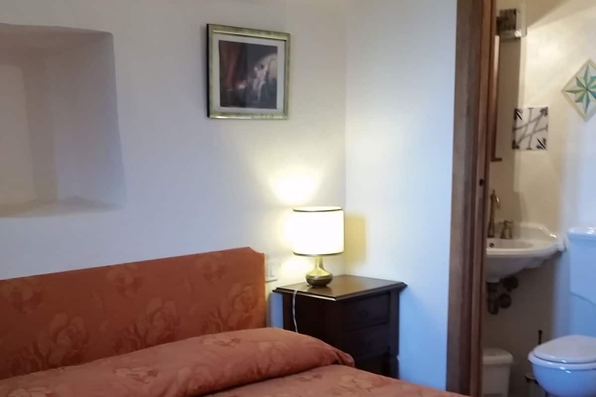 Une chambre à coucher avec un lit, une table de chevet avec une lampe, un tableau encadré au mur et une salle de bain visible à travers une porte ouverte.