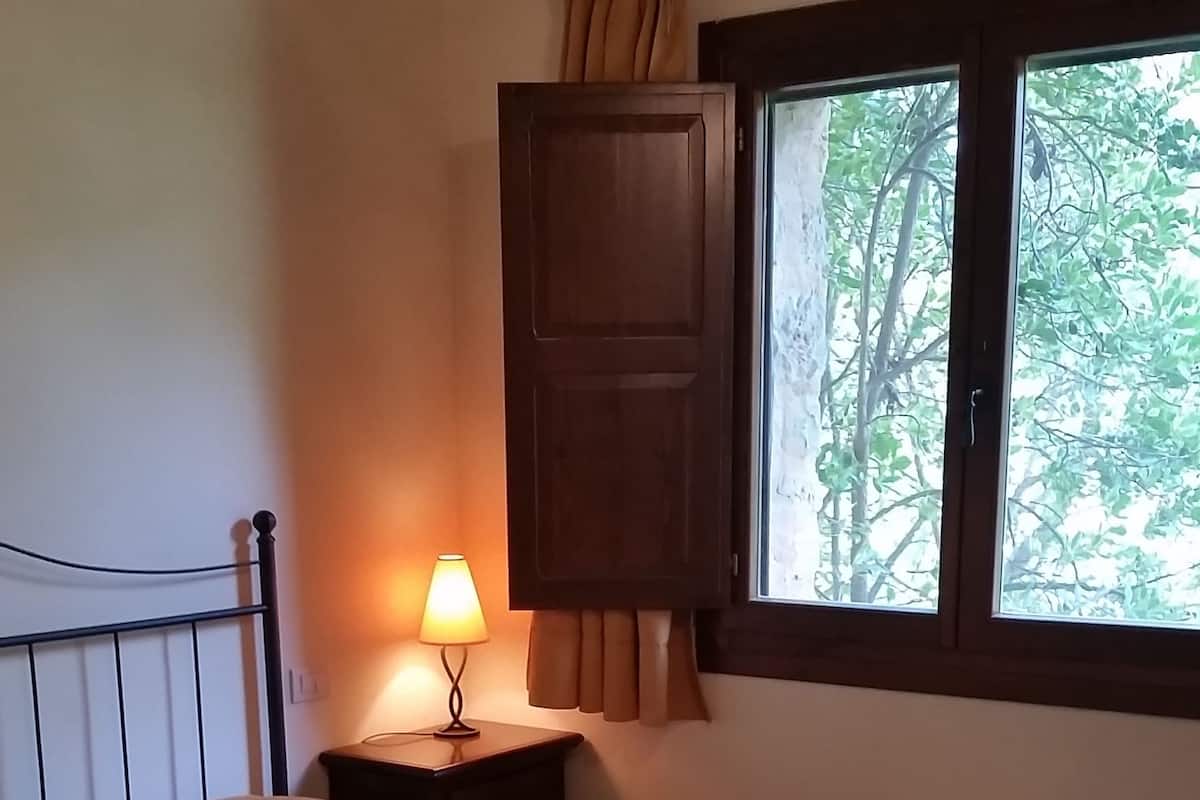Une chambre à coucher avec un lit, une table de chevet avec une lampe, une fenêtre avec des rideaux et une vue sur les arbres à l’extérieur.