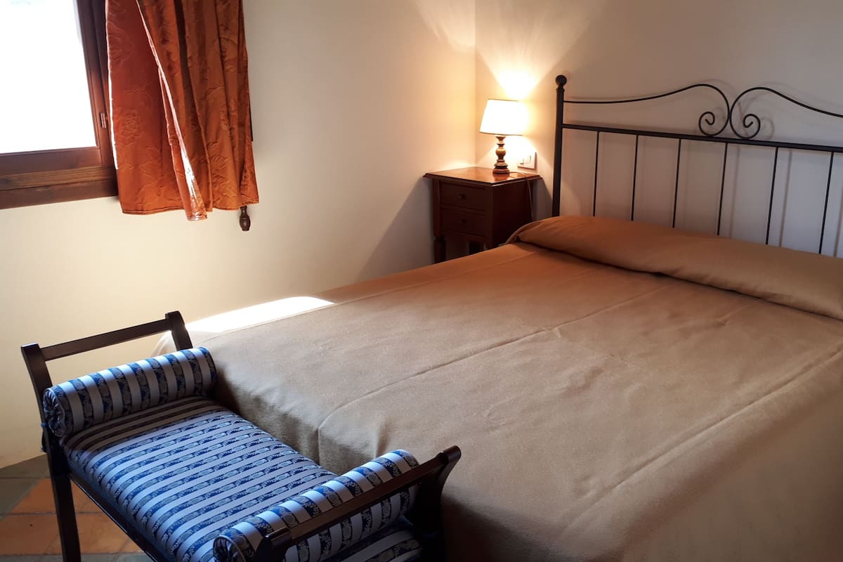 Une chambre à coucher avec un lit, une table de chevet avec une lampe, une chaise et une fenêtre avec des rideaux.