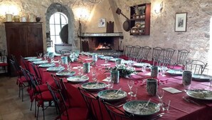 Une longue table à manger, dressée avec des assiettes, des verres et des couverts, entourée de chaises, dans une pièce aux murs de pierre et dotée d’une cheminée.