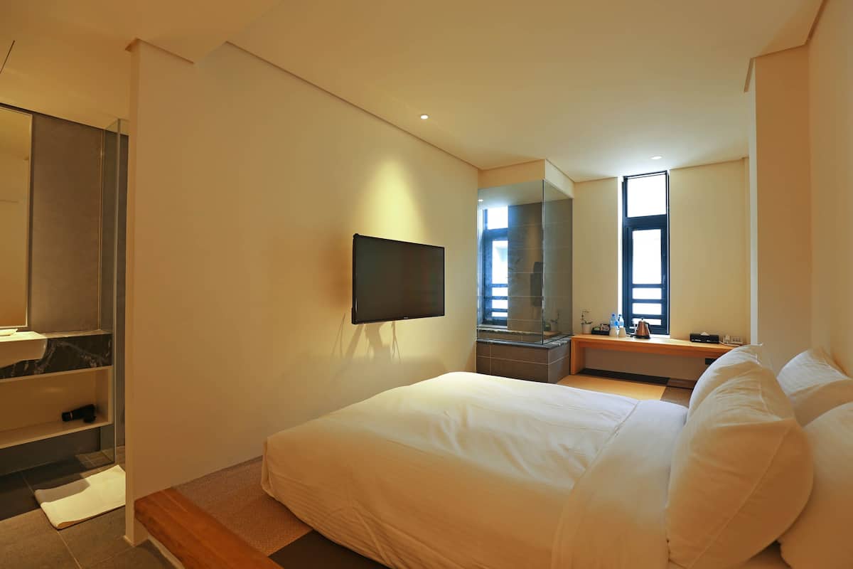 A modern hotel room with a large bed, a flat-screen TV mounted on the wall, a bathroom with a glass shower enclosure, and a large window providing natural light.