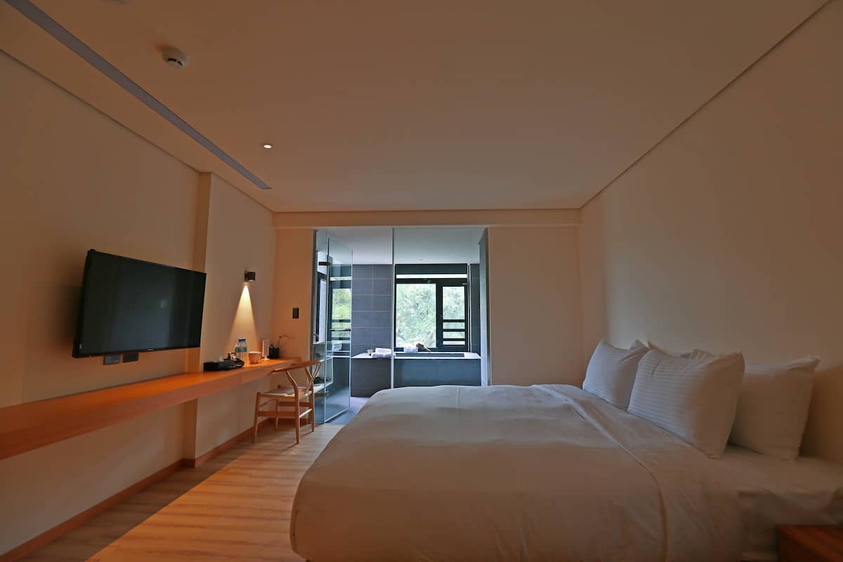 A hotel room with a large bed, a flat-screen TV mounted on the wall, a desk with a chair, and a window offering a view of the outdoors.