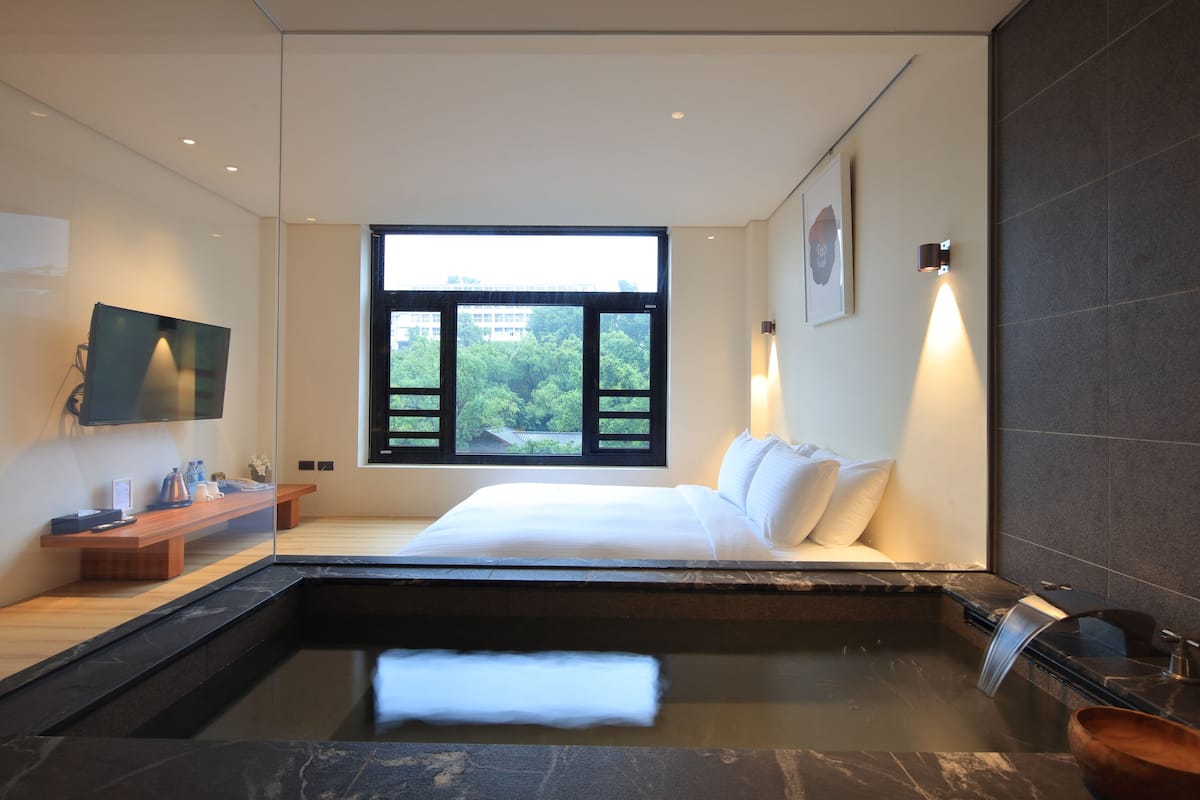 A modern bedroom with a large bed, a small sitting area, a window with a view of greenery, and a built-in hot tub.