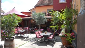 Une terrasse extérieure avec des tables et des chaises, entourée de plantes en pot et agrémentée d’un parasol rouge.