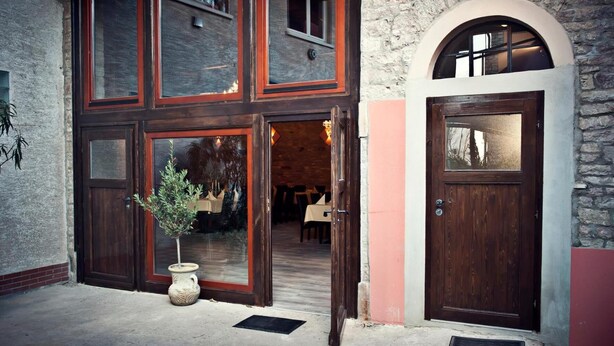 Un bâtiment avec une façade en pierre, une grande porte en bois et un pot de fleurs placé devant l’entrée.